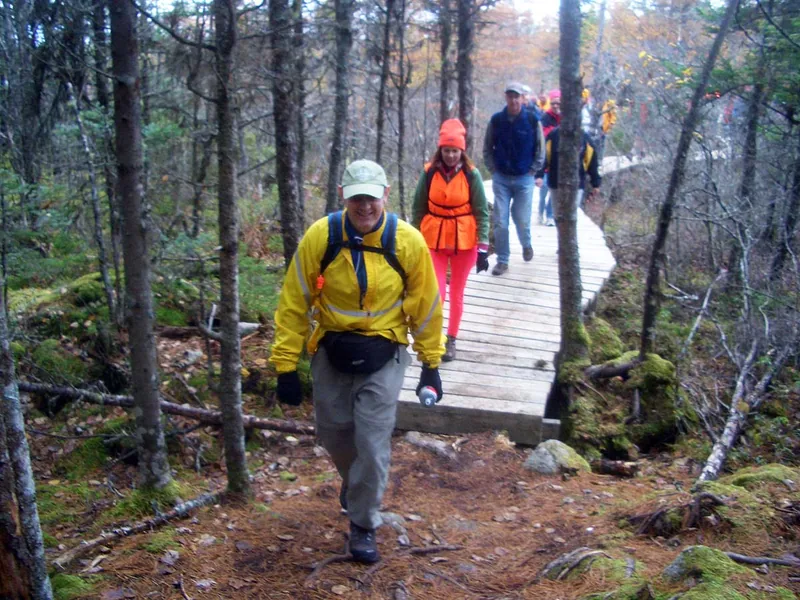A hiker in a red pack crossing a low wooden boardwalk laid over wet ground between mossy boulders.