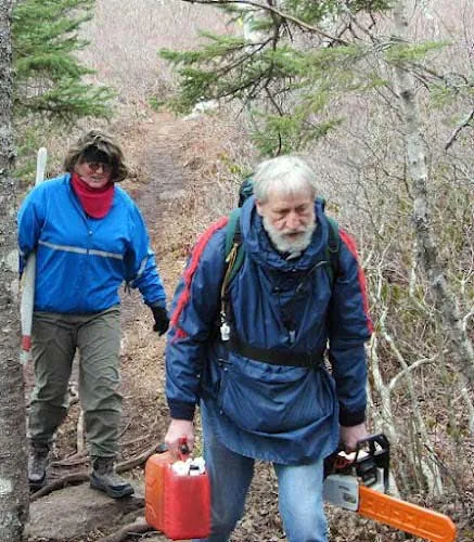 Two WRWEO volunteers on the trail in spring, one carrying a chainsaw and a fuel can, clearing blowdown.